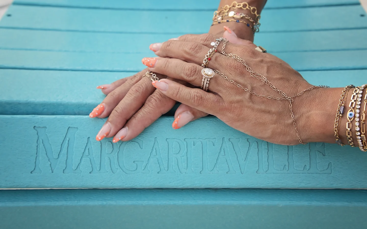 Layered gold permanent bracelets, rings and a hand-chain on hands resting on a turquoise Margaritaville step at Margaritaville Beach Resort Fort Myers Beach